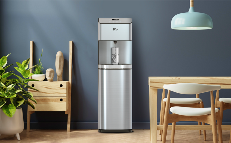 Brio stainless steel water dispenser pouring water into a glass, placed in a modern dining area with wooden furniture, indoor plants, and a light blue pendant lamp against a dark blue wall.